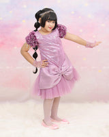 Shimmery Pink Dress With Satin Flower Sleeves Adorned With Pearl Tassels And A Large Bow Accent.
